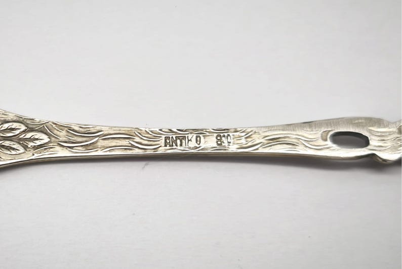 May include: Close-up of a silver antique utensil with intricate leaf and wave patterns. The word "ANTIKO" and the number "800" are stamped on the handle. The utensil has a decorative design.