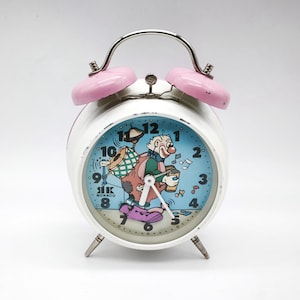 May include: A vintage white alarm clock with pink bells and a silver handle. The clock face features a cartoon clown with a drum, coffee cup, and musical notes on a blue background. The clock reads "SIK MONACO".