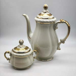 May include: A white porcelain coffee pot and creamer set with gold trim. The coffee pot has a lid with a decorative knob and a curved handle. The creamer has a lid with a decorative knob and two handles.