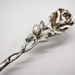 Peut inclure: Une cuill&egrave;re de service argent&eacute;e avec un motif de rose. La cuill&egrave;re pr&eacute;sente une fleur de rose d&eacute;taill&eacute;e en haut, avec des feuilles et une tige torsad&eacute;e. La cuill&egrave;re est probablement en m&eacute;tal et a un aspect vintage ou antique.