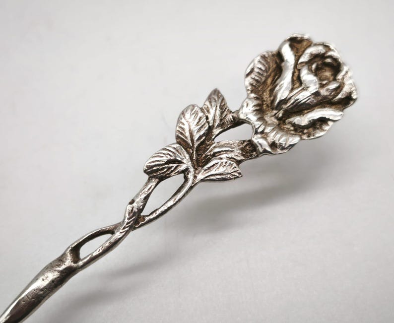 Peut inclure: Une cuill&egrave;re de service argent&eacute;e avec un motif de rose. La cuill&egrave;re pr&eacute;sente une fleur de rose d&eacute;taill&eacute;e en haut, avec des feuilles et une tige torsad&eacute;e. La cuill&egrave;re est probablement en m&eacute;tal et a un aspect vintage ou antique.