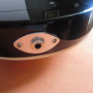 May include: Close-up of a black electric guitar body with a chrome jack plate. The jack plate is oval-shaped and secured with two screws. The guitar's body has a glossy finish and a cream-colored binding.