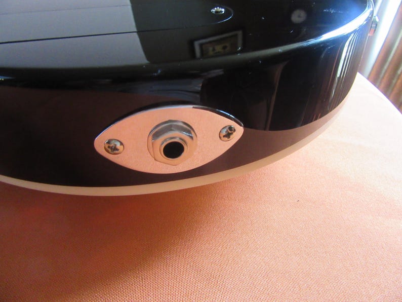 May include: Close-up of a black electric guitar body with a chrome jack plate. The jack plate is oval-shaped and secured with two screws. The guitar's body has a glossy finish and a cream-colored binding.