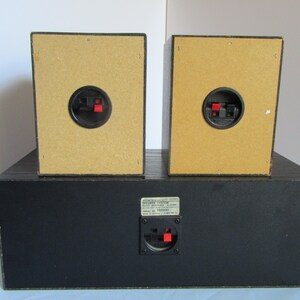 May include: A black speaker system with two smaller speakers on top of a larger base. The speakers have tan backs with black and red connectors. The base has a black finish and a connector with a label.