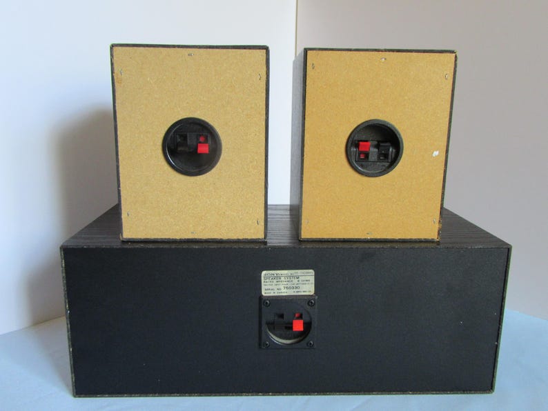 May include: A black speaker system with two smaller speakers on top of a larger base. The speakers have tan backs with black and red connectors. The base has a black finish and a connector with a label.