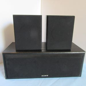 May include: A black Sony home theater speaker system. The system includes a subwoofer and two satellite speakers. The speakers are rectangular and covered in black fabric. The Sony logo is visible on the front of the subwoofer.