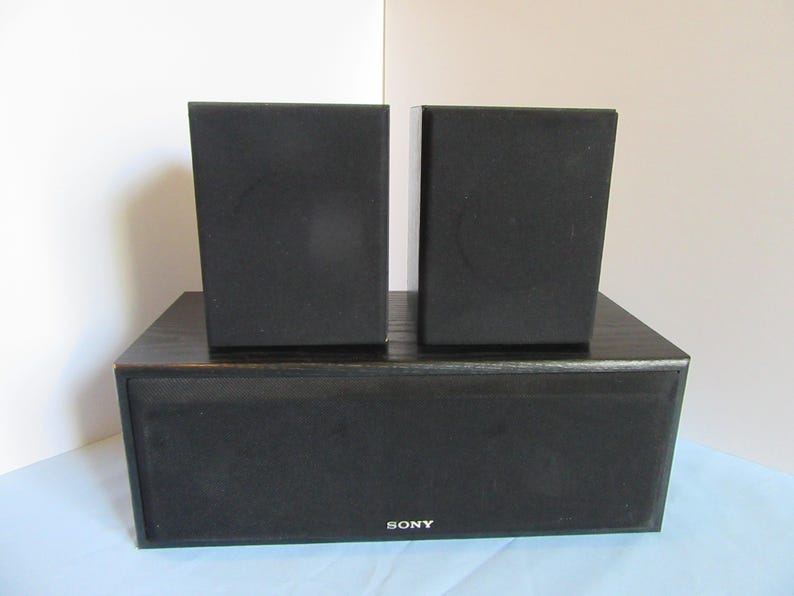 May include: A black Sony home theater speaker system. The system includes a subwoofer and two satellite speakers. The speakers are rectangular and covered in black fabric. The Sony logo is visible on the front of the subwoofer.