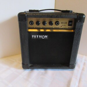 Peut inclure: Amplificateur de guitare Python AG-10 noir avec des boutons et du texte dorés. L'amplificateur a une poignée noire et une grille de haut-parleur avec le mot "PYTHON" en lettres blanches. L'amplificateur est sur une surface blanche.