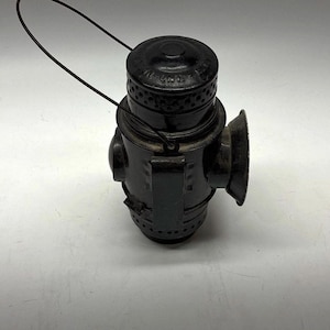 May include: A black metal railroad lantern featuring a cylindrical body, a top handle, and a side-mounted reflector. The lantern has a series of small ventilation holes around the top and bottom sections, with a prominent vertical clip on the side.