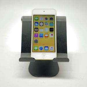 May include: A white iPod Touch with a yellow back casing is positioned vertically on a black metal stand. The screen displays a standard grid of colorful application icons including FaceTime, Calendar, Photos, Camera, Weather, Clock, Maps, and Videos.