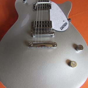 May include: A silver electric guitar with a white pickguard and chrome hardware. The guitar has a classic body shape, visible strings, and tuning knobs. The brand name "Gretsch" is printed on the pickguard.