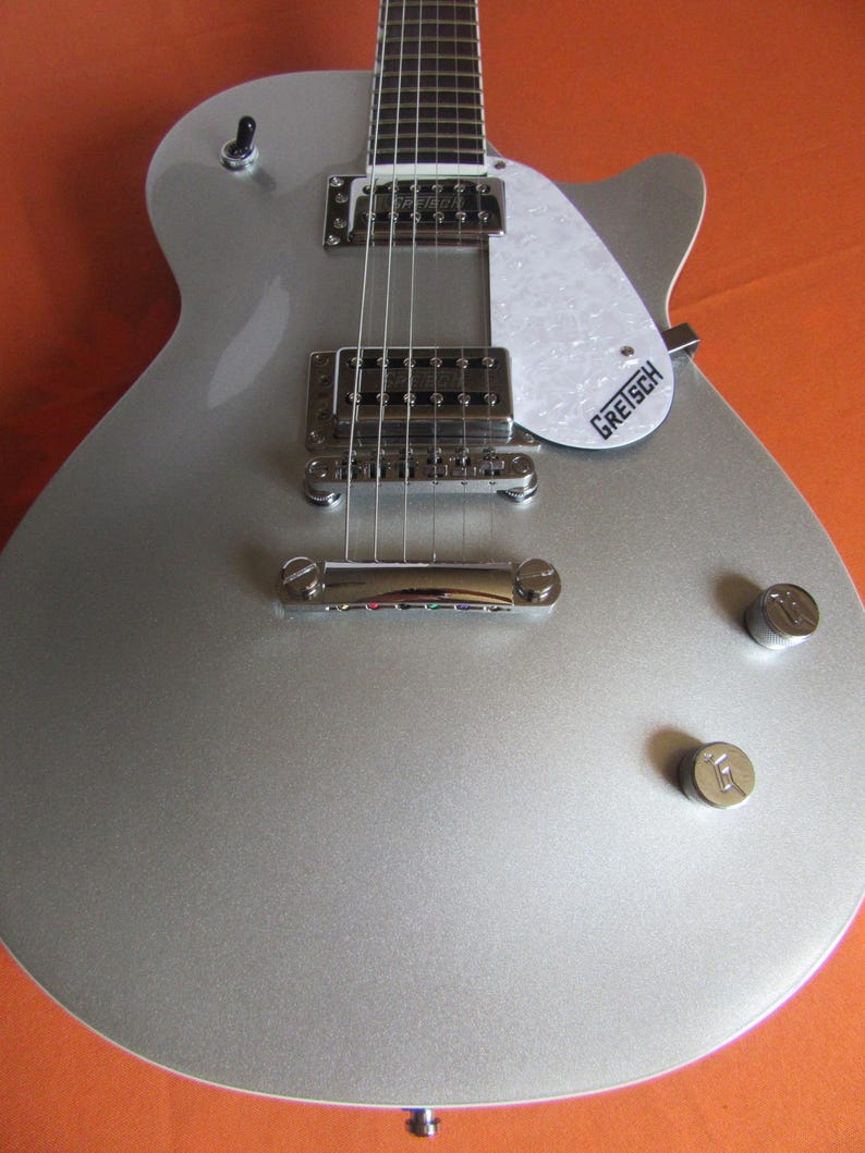 May include: A silver electric guitar with a white pickguard and chrome hardware. The guitar has a classic body shape, visible strings, and tuning knobs. The brand name "Gretsch" is printed on the pickguard.