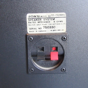 May include: Close-up of a black speaker panel with a white label. The label reads "SONY SPEAKER SYSTEM" with technical specifications. Below is a black input panel with red and black terminals. The speaker is made in Canada.