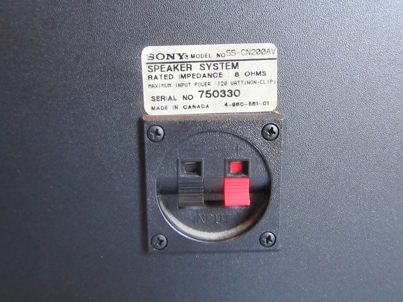 May include: Close-up of a black speaker panel with a white label. The label reads "SONY SPEAKER SYSTEM" with technical specifications. Below is a black input panel with red and black terminals. The speaker is made in Canada.