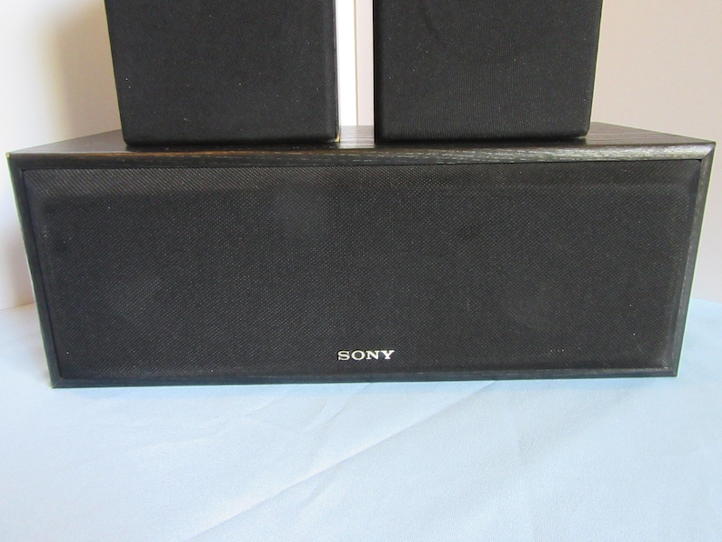 May include: A black Sony speaker system with a rectangular center speaker and two smaller speakers stacked on top. The speakers have a mesh front and the Sony logo is visible.