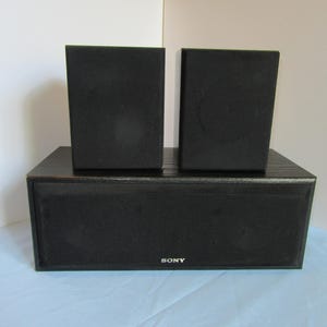 May include: A black Sony speaker system with two smaller speakers on top of a larger rectangular speaker. The speakers are made of a dark material and are set against a white and blue background.