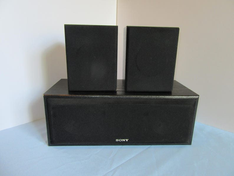 May include: A black Sony speaker system with two smaller speakers on top of a larger rectangular speaker. The speakers are made of a dark material and are set against a white and blue background.