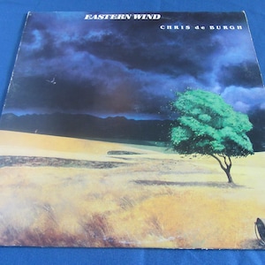 May include: Album cover for "Eastern Wind" by Chris de Burgh. The artwork depicts a landscape with a green tree, a dark, stormy sky, and a bucket. The album title and artist's name are displayed at the top.