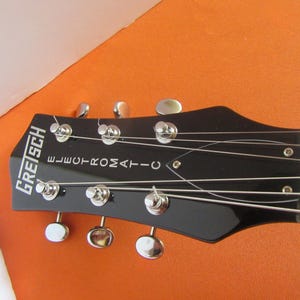 May include: Close-up of a black Gretsch Electromatic guitar headstock. The headstock features silver tuning pegs and the brand name in white lettering. The strings are visible, stretched across the neck.