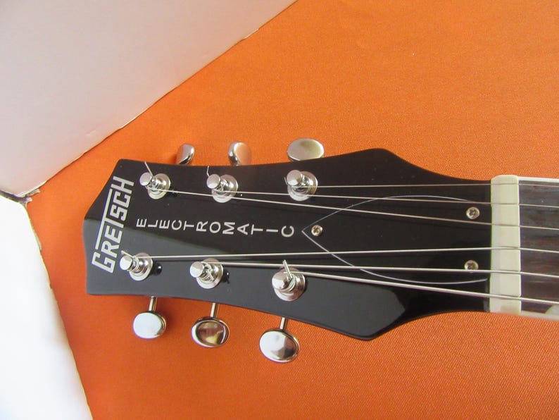May include: Close-up of a black Gretsch Electromatic guitar headstock. The headstock features silver tuning pegs and the brand name in white lettering. The strings are visible, stretched across the neck.