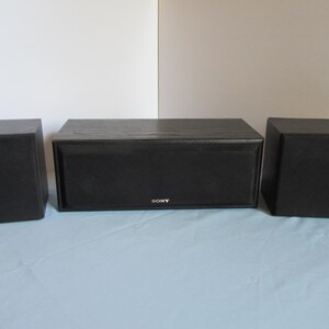 May include: A set of three black Sony speakers. The center speaker is rectangular, with the Sony logo. Two smaller, square speakers flank the center speaker. The speakers are on a light blue surface.
