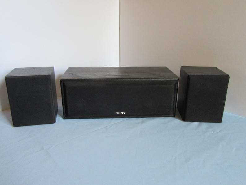 May include: A set of three black Sony speakers. The center speaker is rectangular, with the Sony logo. Two smaller, square speakers flank the center speaker. The speakers are on a light blue surface.