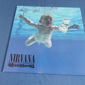 May include: Album cover showing a baby submerged in water, with a dollar bill in front of it. The band name "NIRVANA" and album title "NEVERMIND" are printed in black at the bottom. The background is a blue gradient.