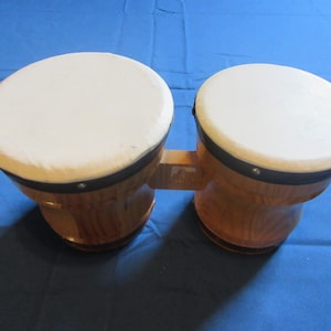 May include: A pair of wooden bongo drums with natural wood grain and black rims. The drums are connected by a wooden bar.