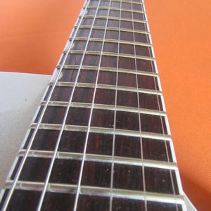 May include: Close-up of a guitar neck with dark brown fretboard and silver frets. The strings are visible, and the neck is angled towards the top right. The background is a solid orange color.