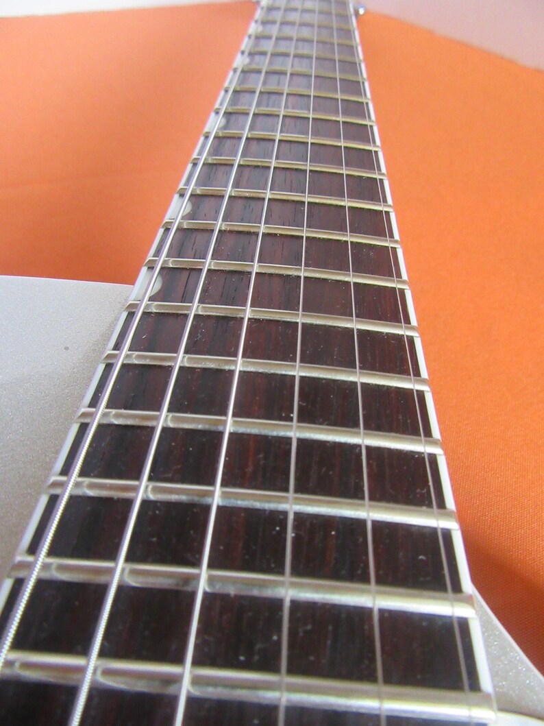May include: Close-up of a guitar neck with dark brown fretboard and silver frets. The strings are visible, and the neck is angled towards the top right. The background is a solid orange color.