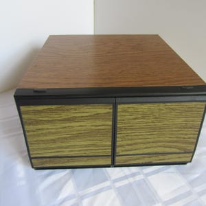 May include: A vintage wooden storage cabinet with a two-door design. The top is a medium brown with a wood grain pattern, while the doors have a lighter, yellowish-green wood grain. The cabinet is trimmed with black accents.