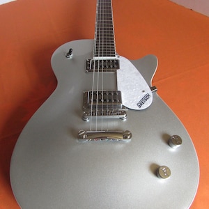 May include: Silver electric guitar with a white pickguard and chrome hardware. The guitar has a classic design with two pickups, volume knobs, and a bridge. The pickguard has the "Gretsch" logo.
