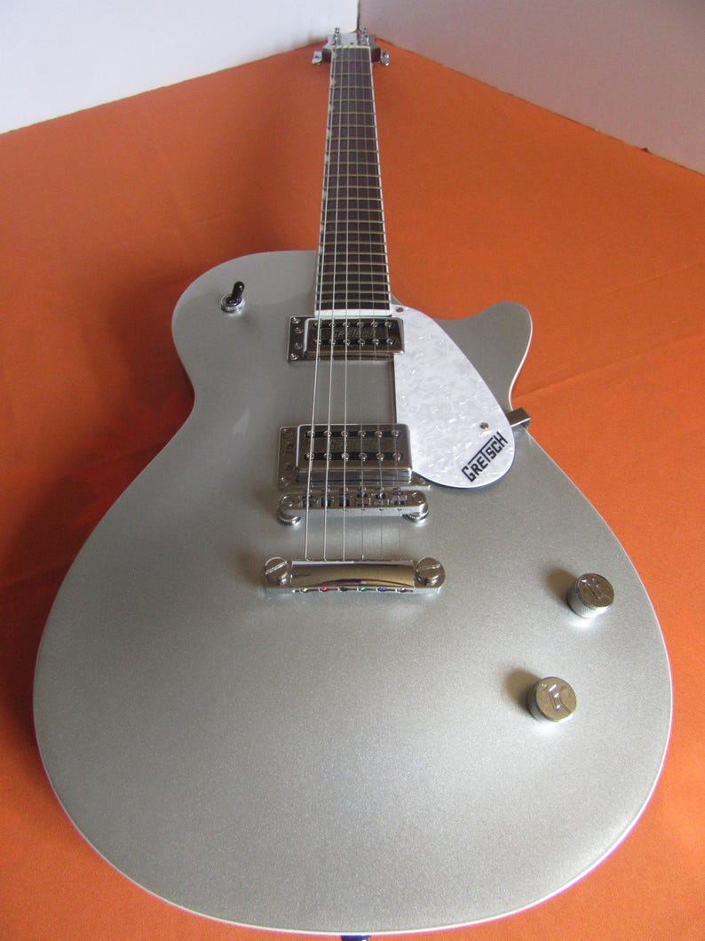 May include: Silver electric guitar with a white pickguard and chrome hardware. The guitar has a classic design with two pickups, volume knobs, and a bridge. The pickguard has the "Gretsch" logo.
