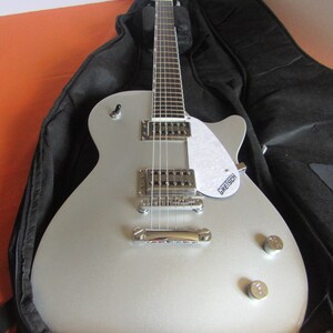 May include: Silver electric guitar with a white pickguard and chrome hardware, resting in a black case. The guitar features a classic design with two control knobs and a bridge. The brand name "Gretsch" is visible on the pickguard.