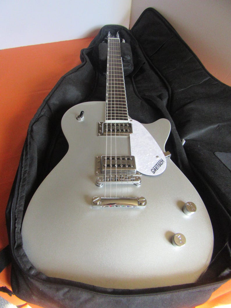 May include: Silver electric guitar with a white pickguard and chrome hardware, resting in a black case. The guitar features a classic design with two control knobs and a bridge. The brand name "Gretsch" is visible on the pickguard.