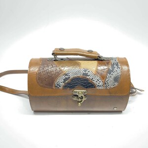 Peut inclure: Un sac à main cylindrique marron doté d'une poignée supérieure et d'une longue bandoulière. L'extérieur présente un motif patchwork avec du cuir marron lisse, des panneaux texturés façon crocodile et des accents à motif peau de serpent.
