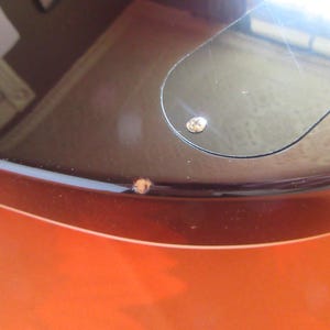 May include: Close-up of a guitar body with a glossy finish. The body transitions from a vibrant orange to a dark brown, with a black pickguard and a visible screw. The image highlights the instrument's curves and color gradient.