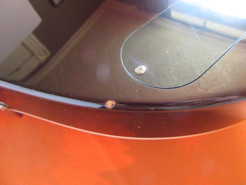 May include: Close-up of a guitar body with a glossy finish. The body transitions from a vibrant orange to a dark brown, with a black pickguard and a visible screw. The image highlights the instrument's curves and color gradient.