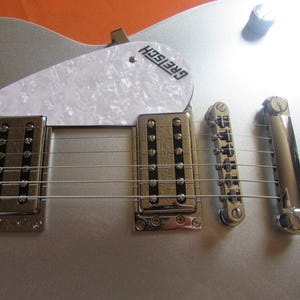 May include: Close-up of a silver Gretsch guitar. The guitar features a white pickguard with the Gretsch logo, chrome hardware, and six strings. The body is a metallic silver color.