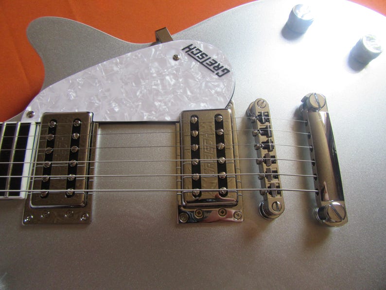 May include: Close-up of a silver Gretsch guitar. The guitar features a white pickguard with the Gretsch logo, chrome hardware, and six strings. The body is a metallic silver color.