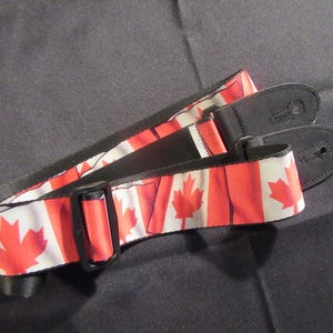 May include: A black guitar strap with a Canadian flag design. The strap is made of fabric and has a black leather end piece.