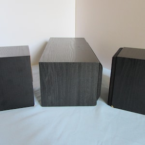 May include: Three black wooden speakers with a dark wood grain finish. The speakers are rectangular, with a slightly angled top. One speaker is centered, and two are on either side.
