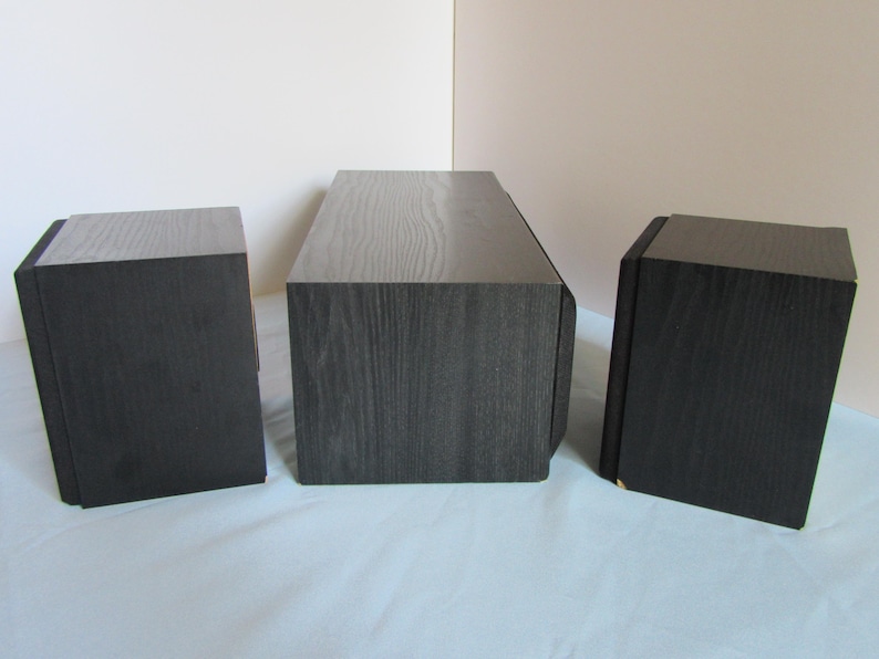 May include: Three black wooden speakers with a dark wood grain finish. The speakers are rectangular, with a slightly angled top. One speaker is centered, and two are on either side.