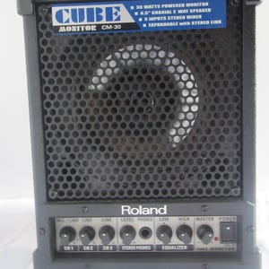 May include: Black Roland Cube Monitor CM-30 speaker. The speaker has a black metal grill covering the speaker. The control panel has knobs for mic/line, line, level phones, low, high, master, and power. The speaker has the text "CUBE MONITOR CM-30".