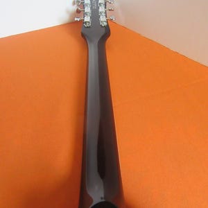 May include: A close-up of the headstock and neck of a black electric guitar. The headstock features silver tuning pegs and a brand name. The neck is glossy black, contrasting with the orange and white background.