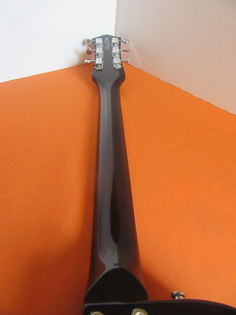 May include: A close-up of the headstock and neck of a black electric guitar. The headstock features silver tuning pegs and a brand name. The neck is glossy black, contrasting with the orange and white background.