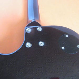 May include: Close-up of the back of a black electric guitar. The guitar has a blue neck and headstock. The body is textured with a repeating pattern. Silver screws secure the neck and a black plate to the body.