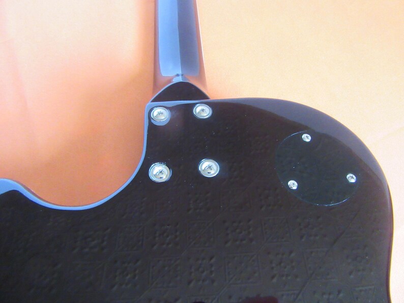 May include: Close-up of the back of a black electric guitar. The guitar has a blue neck and headstock. The body is textured with a repeating pattern. Silver screws secure the neck and a black plate to the body.