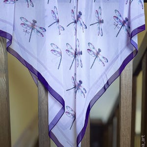 May include: A square scarf featuring a dragonfly design in purple and blue hues on a white backdrop. The scarf has a dark purple border and is draped over a wooden banister.