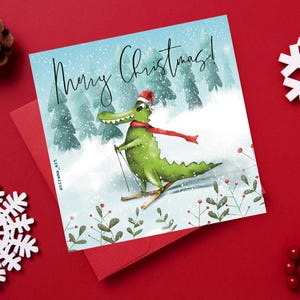 May include: A Christmas card featuring a green alligator wearing a red Santa hat and scarf, skiing down a snowy hill. The card reads "Merry Christmas!"